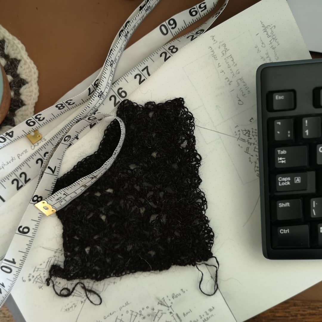 a swatch and tape measure lie on an open design journal, the edge of a crochet coaster and a computer keyboard can also be seen in shot.