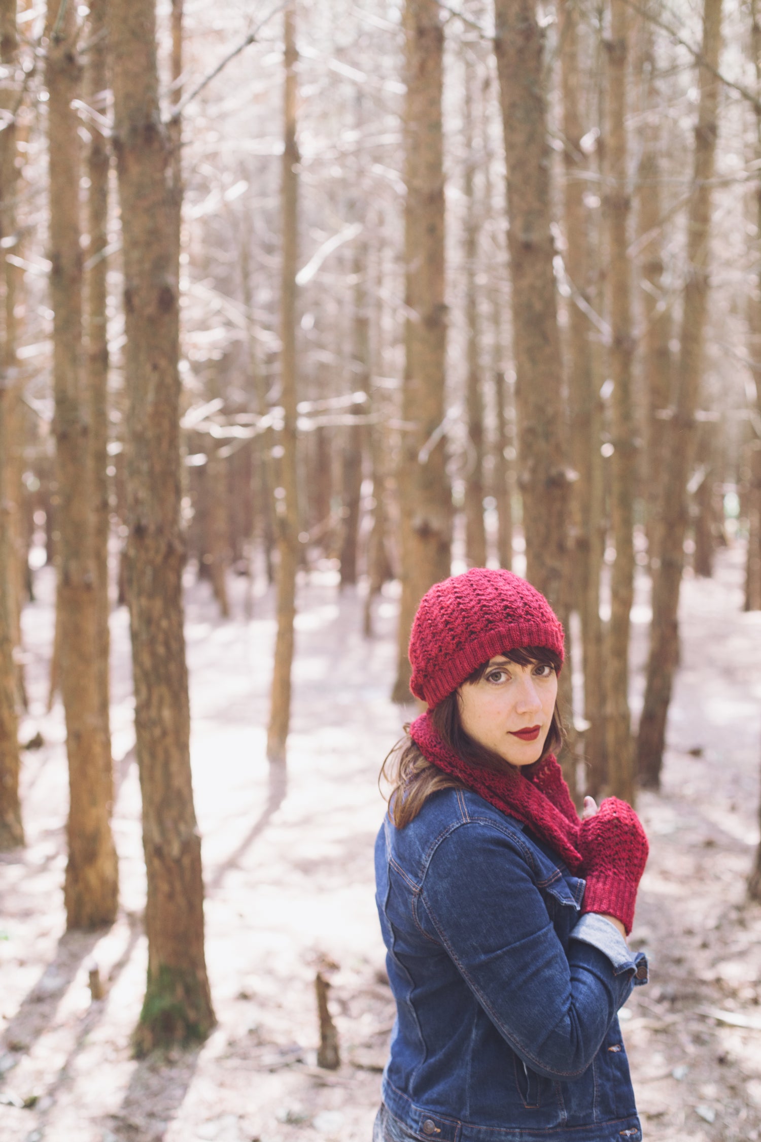 a woman stands in snowy woods in a denim jacket and bright red hat, cowl and mitts set, she turns to look at the camera