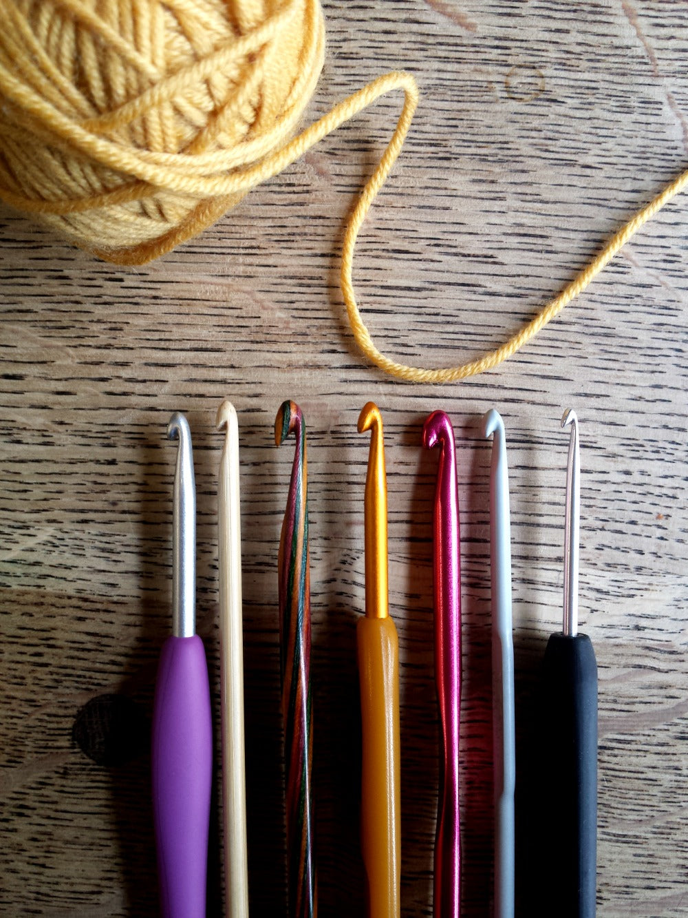 a range of crochet hooks and a yellow ball of yarn sit on a wooden background